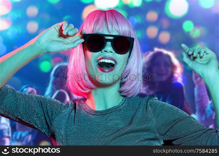 nightlife, entertainment and people concept - happy young woman wearing pink wig and black sunglasses dancing at nightclub over lights background. woman in wig and sunglasses dancing at nightclub