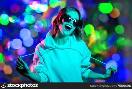 nightlife, fashion and people concept - happy young woman in black sunglasses wearing hoodie over night lights background. woman in hoodie and sunglasses over night lights