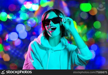 nightlife, fashion and people concept - happy young woman in black sunglasses wearing hoodie over night lights background. woman in hoodie with sunglasses over night lights