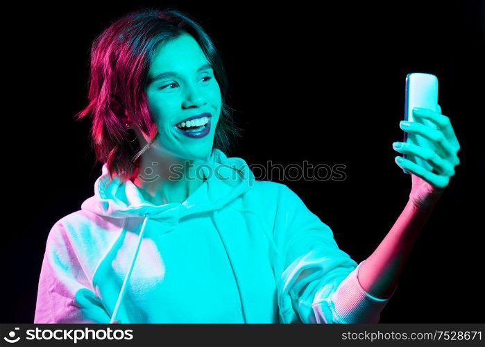 nightlife, fashion and people concept - happy young woman wearing hoodie taking selfie by smartphone in neon lights over black background. woman taking selfie by smartphone in neon lights