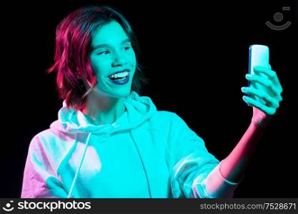 nightlife, fashion and people concept - happy young woman wearing hoodie taking selfie by smartphone in neon lights over black background. woman taking selfie by smartphone in neon lights