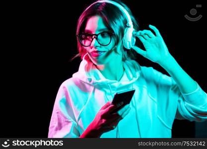 nightlife, fashion and people concept - happy young woman wearing hoodie with smartphone and headphones in neon lights over black background. woman in headphones with smartphone in neon lights