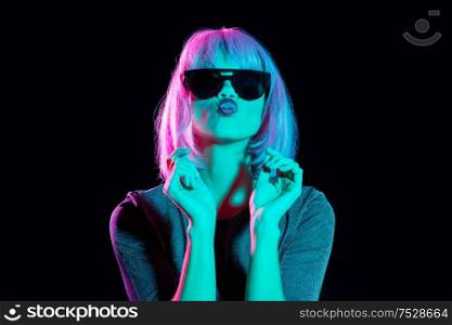nightlife, fashion and people concept - happy young woman wearing pink wig and black sunglasses in neon ultra violet light sending air kiss over black background. woman in wig and black sunglasses sending air kiss