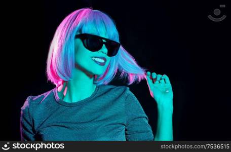 nightlife, fashion and people concept - happy young woman wearing pink wig and black sunglasses in neon ultra violet light over black background. happy woman in pink wig and black sunglasses