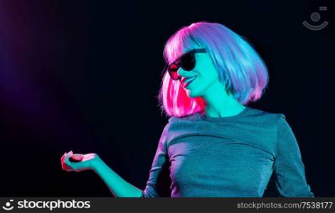 nightlife, fashion and people concept - happy young woman wearing pink wig and black sunglasses in neon ultra violet light dancing over black background. happy woman in pink wig and sunglasses dancing