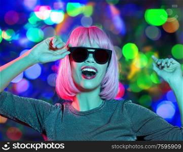 nightlife, fashion and people concept - happy young woman wearing pink wig and black sunglasses in neon ultraviolet light dancing over festive lights background. happy woman in pink wig and sunglasses dancing