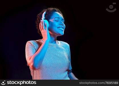 nightlife, technology and people concept - happy young african american woman in headphones listening to music and dancing in neon lights over black background. woman in headphones listening to music and dancing