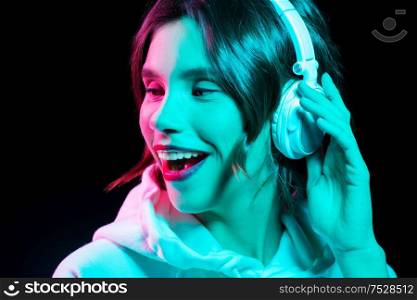 nightlife, technology and people concept - happy young woman in headphones wearing hoodie listening to music in neon lights over black background. woman in headphones listening to music in neon