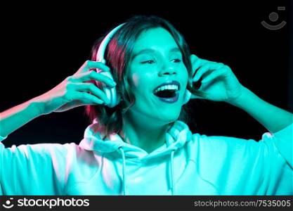 nightlife, technology and people concept - happy young woman in headphones wearing hoodie listening to music in neon lights over black background. woman in headphones listening to music in neon
