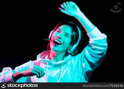 nightlife, technology and people concept - happy young woman in headphones wearing hoodie listening to music and dancing in neon lights over black background. woman in headphones listening to music and dancing