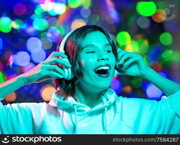 nightlife, technology and people concept - happy young woman in headphones wearing hoodie listening to music over night lights background. woman in headphones listening to music over lights