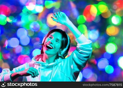 nightlife, technology and people concept - happy young woman in headphones wearing hoodie listening to music and dancing over night lights background. woman in headphones dancing over night lights