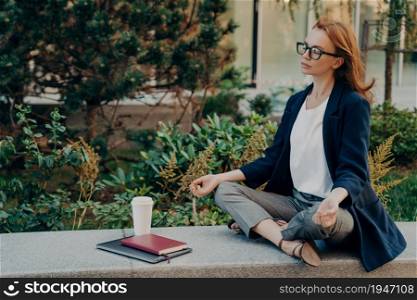 No stress. Calm red-haired woman with closed eyes relaxing while working remotely on laptop outdoors, mindful young business woman meditating with closed eyes, doing breathing yoga exercises outside. Calm red-haired woman relaxing meditating while working remotely on laptop outdoors