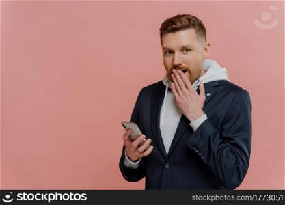 No way. Amazed young man with red hair in casual wear staring at camera with stunned expression, reading unexpected news on smartphone while standing isolated over pastel pink background. Young man receiving shocking message on smartphone