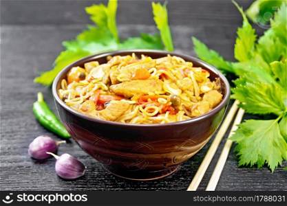 Noodles with cabbage, chicken breast, sweet red peppers, onions and peas, seasoned with soy sauce in a bowl, garlic and celery leaves on dark wooden board background