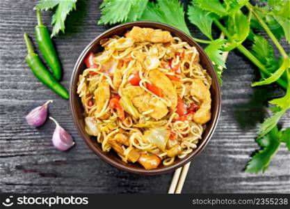 Noodles with cabbage, chicken breast, sweet red peppers, onions and peas seasoned with soy sauce in a bowl, garlic and celery leaves on wooden board background from above