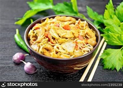 Noodles with cabbage, chicken breast, sweet red peppers, onions and peas, seasoned with soy sauce in a bowl, garlic and green celery leaves on wooden board background