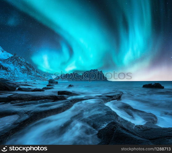 Northern lights above snowy mountains and sandy beach with stones. Aurora borealis in Lofoten islands, Norway. Starry sky with polar lights. Night winter landscape with aurora, sea with blurred water