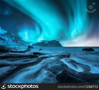 Northern lights above snowy mountains and sandy beach with stones. Aurora borealis in Lofoten islands, Norway. Starry sky with polar lights. Night winter landscape with aurora, sea with blurred water
