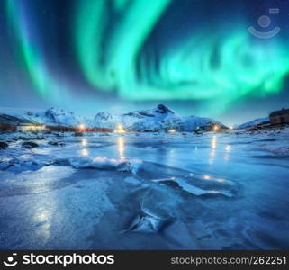 Northern lights above snowy mountains, frozen sea coast and houses in Lofoten islands, Norway. Aurora borealis and small village. Winter landscape with polar lights, rorbu, ice. Aurora over rocks. Northern lights above snowy mountains, frozen sea coast