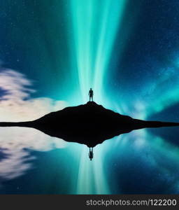 Northern lights and silhouette of standing man in the hill in Norway. Aurora borealis and man. Stars and green polar lights. Night landscape with aurora, lake, sky reflection in water. Travel. Concept. Northern lights and silhouette of standing man in the hill