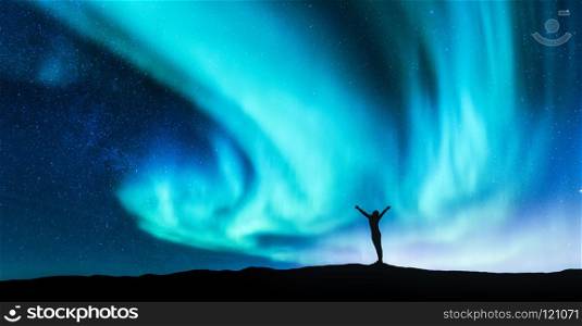 Northern lights and silhouette of standing woman with raised up arms on the hill in Norway. Aurora borealis and happy girl. Starry sky, green polar lights. Night landscape. Travel background. Concept. Northern lights and silhouette of a woman with raised up arms