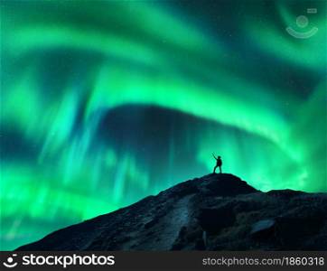 Northern lights and silhouette of standing woman with raised up arms on the mountain. Aurora borealis and happy girl. Sky with stars and green polar lights. Night landscape with aurora. Concept