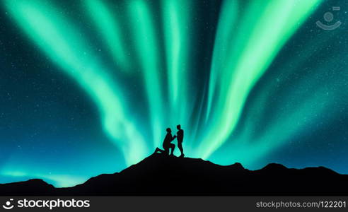 Northern lights and silhouettes of a man making marriage proposal to his girlfriend on the hill. Landscape with night starry sky, aurora borealis, couple. People. Lovers and polar lights. Travel. Northern lights and silhouettes of a couple