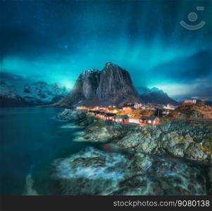 Northern lights in Hamnoy, Lofoten Islands, Norway. Starry sky and Aurora borealis, snowy mountains, sea coast, rorbuer, rocks and stones in water, city lights at winter night. Bright polar lights