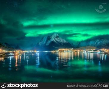Northern lights in Reine, Lofoten Islands, Norway. Starry sky and Aurora borealis, snowy mountains, sea coast, houses, rocks, reflection in water, city lights at winter night. Bright polar lights