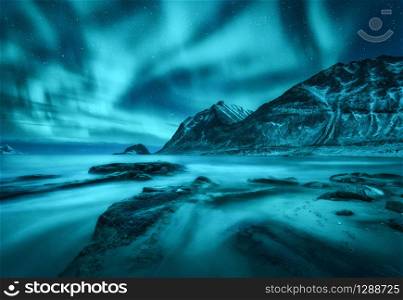 Northern lights over snowy mountains, sea coast with sandy beach and stones in blurred water in Lofoten islands, Norway. Aurora borealis. Winter landscape with polar lights. Starry sky with aurora