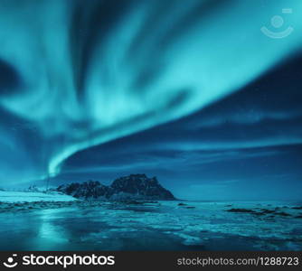 Northern lights over the snowy mountains and sandy beach in winter. Aurora borealis in Lofoten islands, Norway. Starry sky with polar lights. Night landscape with aurora, frozen sea coast, blue sky