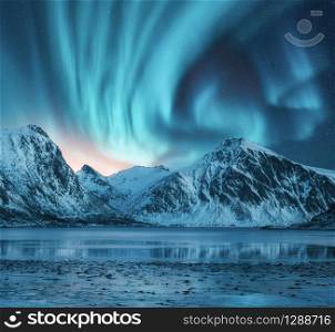Northern lights over the snowy mountains, coast of the lake and reflection in water. Aurora borealis and snow covered rocks. Winter landscape with polar lights, fjord. Starry sky with bright aurora