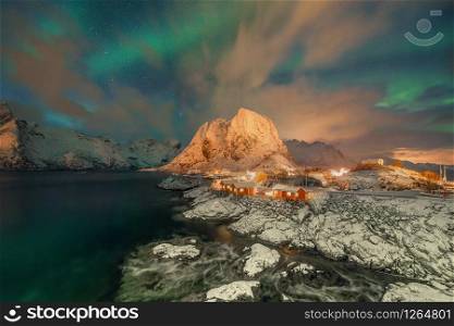 Norwegian fishing village with northern lights and stars in Hamnoy City, Lofoten islands, Norway, Europe. White snowy mountain hills and trees at night, nature landscape background in winter season.