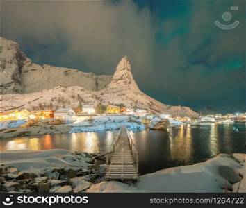 Norwegian fishing village with northern lights and stars in Reine City, Lofoten islands, Norway, Europe. White snowy mountain hills and trees at night, nature landscape background in winter season.