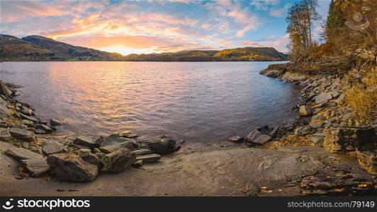 Norwegian fjord landscape with sunset at autumn time. Colorful sky, pastel water and rocky coast with golden trees.