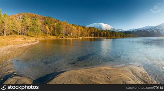 Norwegian landscape with lake, autumn forest and distant snow-covered mountains