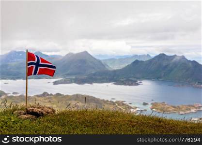 Norwegian national flag waiving in the wind on the top of Nonstinden peak with fjord in the background, Ballstad, Vestvagoy Municipality, Nordland county, Norway