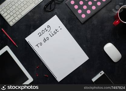 Note book, smart phone and other supplies with cup of coffee with text 2019 to do list . Wooden black office desk table with top view angle .
