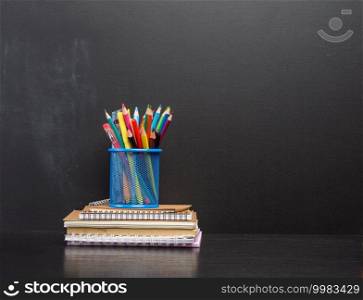 notebook, blue stationery glass with multi-colored wooden pencils on the background of an empty black chalk board, back to school concept