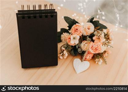 notebook with black sheets with copy space, on a wooden table with a bouquet of flowers and a white heart and bokeh lights around it.