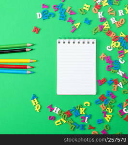 notebook with empty white sheets and multicolored wooden pencils on a green background, top view