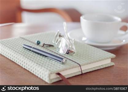 Notebook with pen, eyeglasses and white coffee cup on wooden table, stock photo
