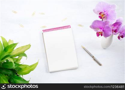 Notepad mockup with green plant leaves, pen, golden paperclip and pink orchid on the white table