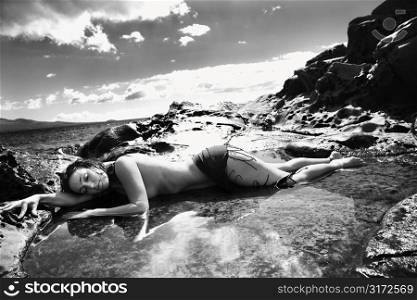 Nude Caucasian mid adult woman lying in tidal pool at Maui coast with eyes closed and head on arm.