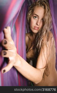 Nude Caucasian woman looking at viewer and reaching out through sheets of colorful fabric.