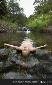 Nude Caucasian young adult woman laying in freshwater stream laughing with head back and arms out.