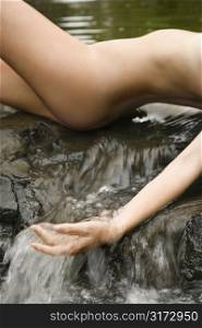 Nude Caucasian young adult woman laying in freshwater stream running water over hands.