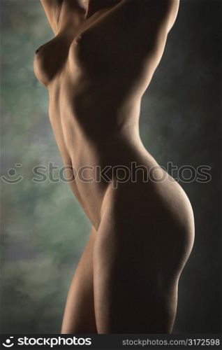 Nude Hispanic mid adult woman standing with arms raised above head.