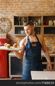 Nude husband in apron holds tray with sweet dessert on the kitchen. Naked male person preparing breakfast at home, food preparation without clothes